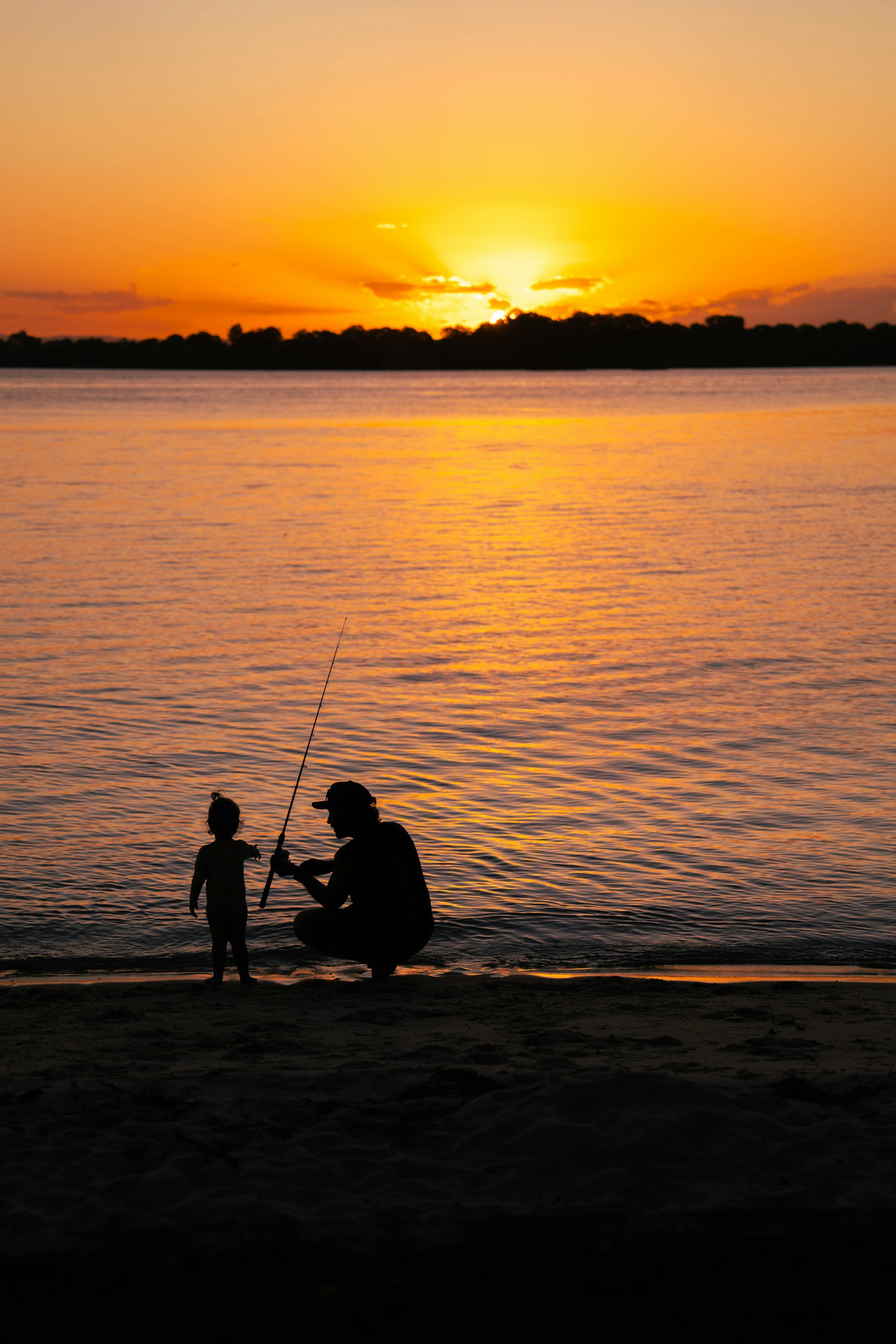 A father helping his toddler hold a fishing rod on the beach at sunset